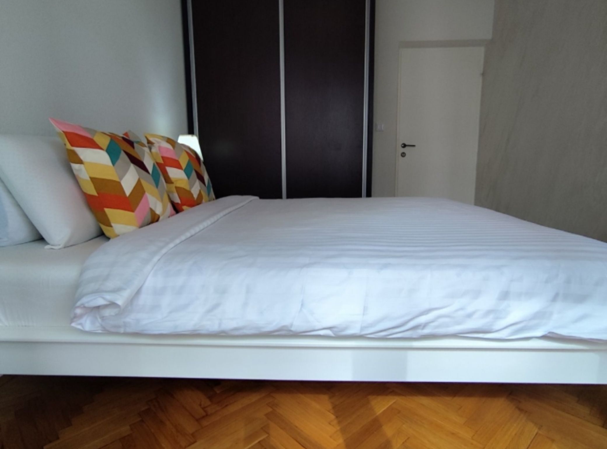 Ein ordentlich bezogenes Bett mit weißer Bettwäsche und farbenfrohen geometrischen Kissen, ein Holzboden mit Fischgrätmuster und ein dunkel gefärbter Kleiderschrank mit Tür.
