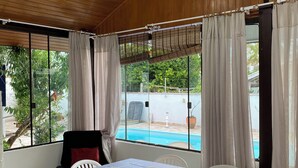 Une pièce avec un plafond en bois, une table recouverte d’une nappe à carreaux, des chaises en plastique blanches et une vue sur une piscine à l’extérieur.
