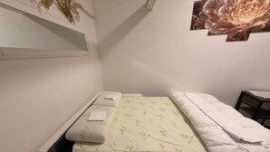 Une petite chambre avec un lit simple, un matelas à motifs de bambou, une table de chevet et une étagère murale agrémentée d’objets décoratifs.