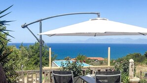 A patio with a table and chairs, a white umbrella, and a view of the sea and mountains.