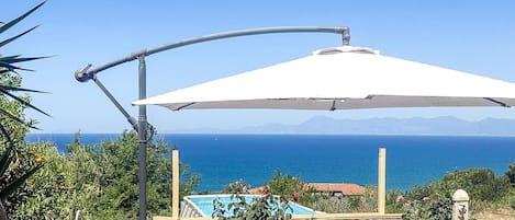 Une terrasse avec une table et des chaises, un parasol blanc, et une vue sur la mer et les montagnes.