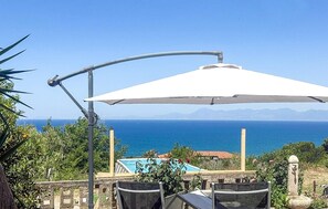 Un pati amb taula i cadires, un parasol blanc i vista al mar i a les muntanyes.
