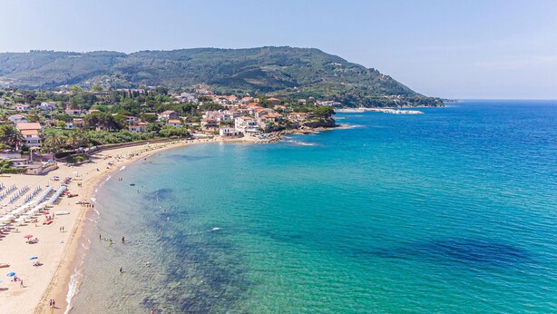 A coastal town with a beach, clear blue waters, and a hillside with buildings.