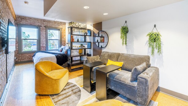 A modern living room with a sofa, armchair, and a coffee table. There is a bookshelf and a hanging plant. - Urban Brick Loft Studio • Ride to Trails • Walk to Dining, Shops & Nightlife (Rogers)