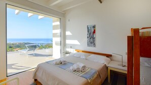 A bedroom with a large window, a bed with rolled towels, a bedside table, and a painting on the wall.