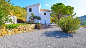 Un edificio blanco con muro de piedra y camino de entrada de grava, rodeado de follaje y árboles.