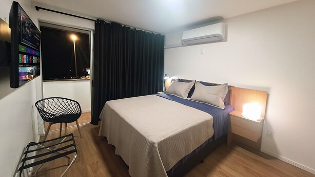 A hotel room with a bed, a chair, a window with curtains, an air conditioning unit, and a wall-mounted TV. - Studio apartment with movement, elevator, and open balcony in Asa Norte. (Brasília)