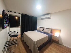A hotel room with a bed, a chair, a window with curtains, an air conditioning unit, and a wall-mounted TV. - Studio apartment with an elevator and an open balcony in Asa Norte. (Brasília)