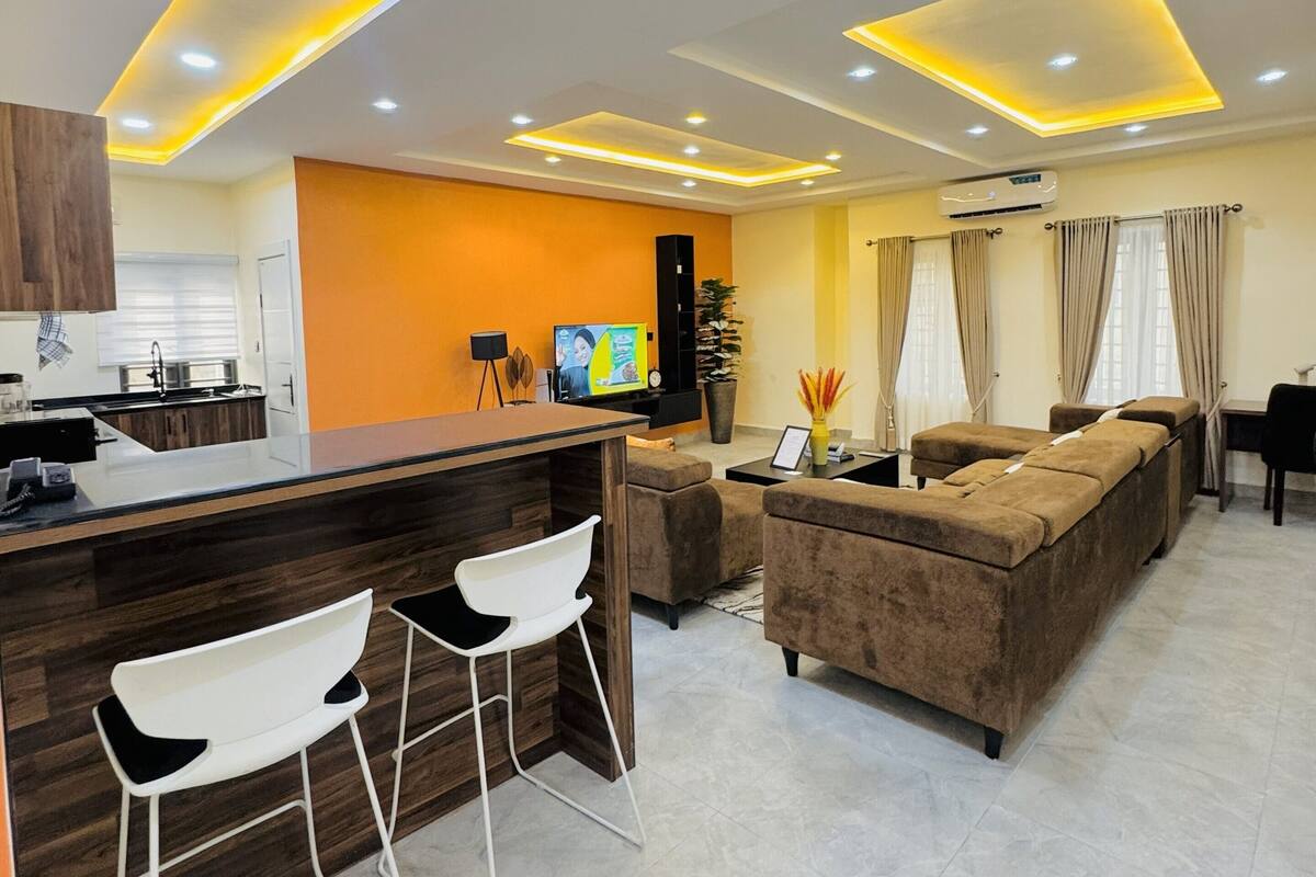 A modern living room with a kitchen area, featuring a bar with stools, a sofa, and a television.