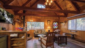 A cozy cabin interior with wooden walls, a dining area, a sofa, a rocking chair, and a large window. - Cozy country cabin in Big Bear perfect for your getaway. Pets wifi. New spa. (Big Bear)