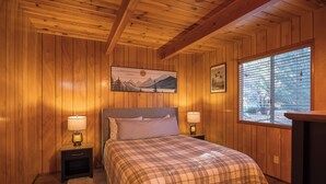 A wooden-paneled bedroom with a bed, two bedside lamps, a painting, and a window with blinds. - Cozy country cabin in Big Bear perfect for your getaway. Pets wifi. New spa. (Big Bear)
