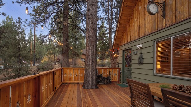 A wooden deck with a dining table and chairs, surrounded by trees and a cabin. - Cozy country cabin in Big Bear perfect for your getaway. Pets wifi. New spa. (Big Bear)