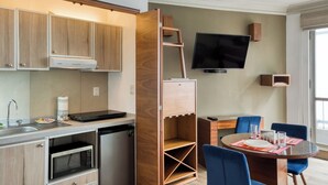 Una cocina funcional con armarios de madera, microondas integrado y lavavajillas. Un televisor de pantalla plana está montado en la pared sobre una zona de comedor con mesa de vidrio y sillas azules.