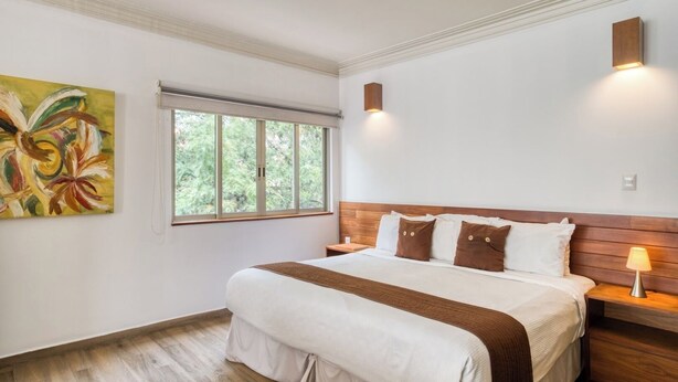 Un dormitorio con una cama grande, un ventanal con vistas a árboles, un cuadro en la pared y una mesita de noche con una lámpara.
