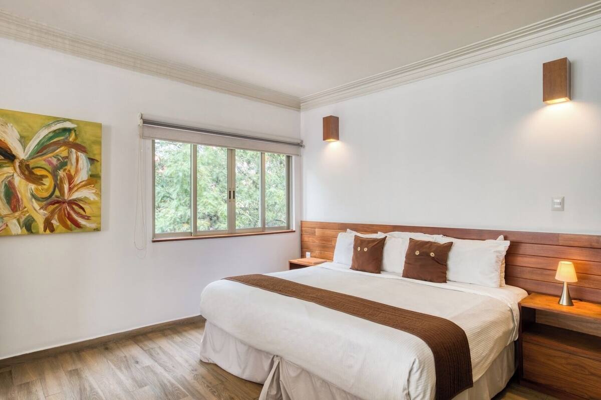 A bedroom with a large bed, a window with a view of trees, a painting on the wall, and a nightstand with a lamp.