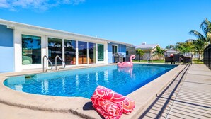 A swimming pool with a pink flamingo float, a covered patio area, and a modern building with large windows. - Clover Vacation Homes: Snead Island: Luxury 4 BR, Heated Pool, Beaches (Palmetto)