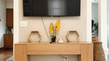 Un salon moderne avec un grand téléviseur à écran plat fixé au mur, une console en bois sur laquelle est posé un vase rempli de fleurs jaunes, et un panier tressé sur le sol.