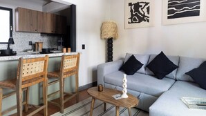 A modern living room with a grey sofa, two wooden chairs, a small wooden table, and a kitchen in the background. - Baranda OCHO By Smart W (Mexico City)