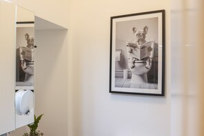 Une photographie encadrée représentant une statue lisant un journal, un porte-rouleau de papier toilette et une plante dans une salle de bain.