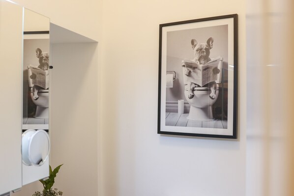 A framed photograph of a statue reading a newspaper, a toilet paper holder, and a plant in a bathroom.