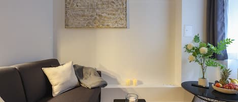 Una sala moderna con un sofá oscuro, un candelabro de vidrio, una mesa redonda con un frutero y un adorno en la pared.