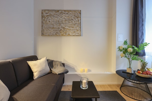 Un salon moderne avec un canapé foncé, un porte-bougie en verre, une table ronde sur laquelle se trouve un saladier de fruits et un élément décoratif mural.