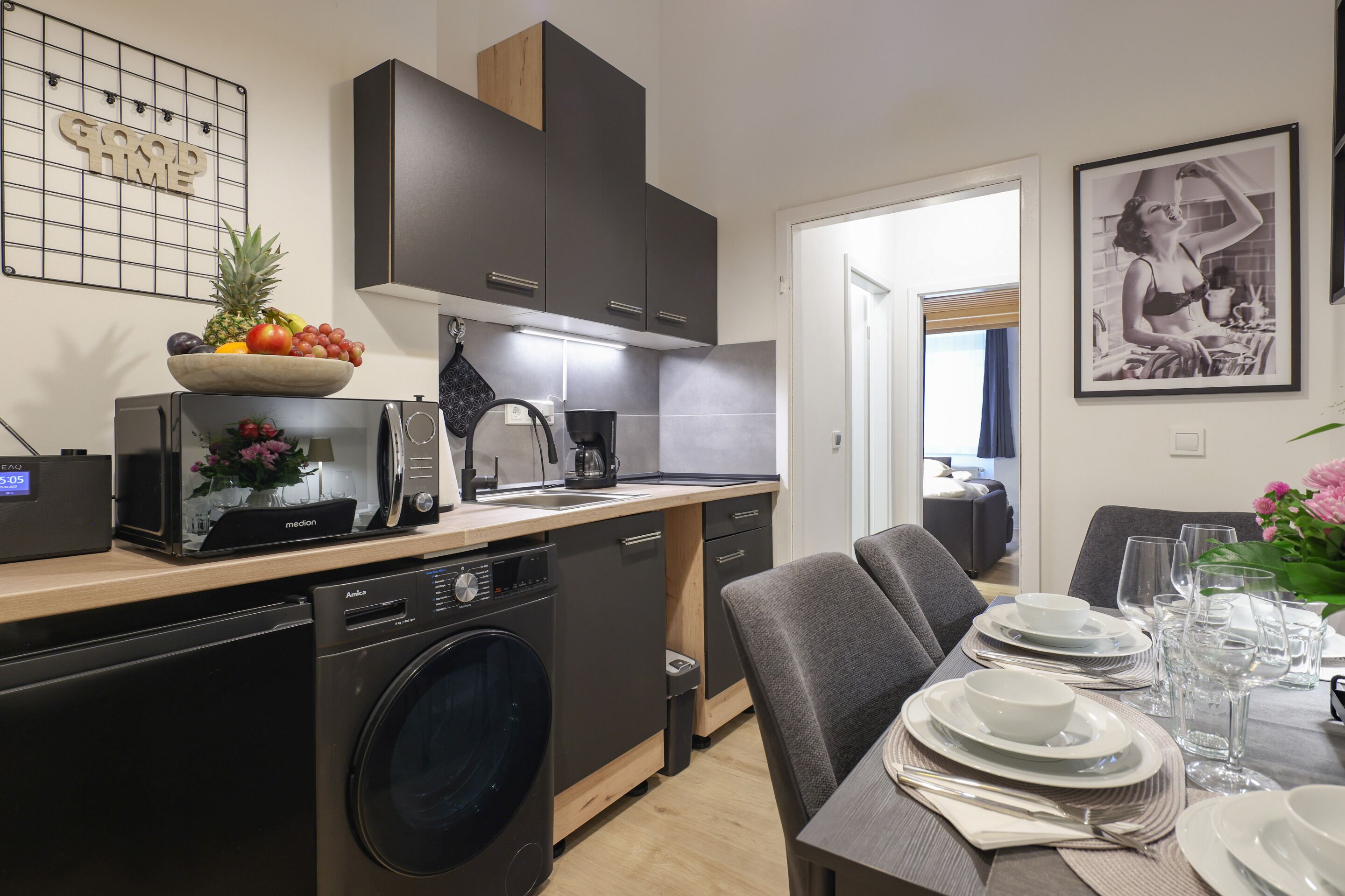 A modern kitchen with a dishwasher, microwave, and coffee maker. There is a dining area with a table set for two, and a framed picture on the wall.