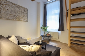 Ein modernes Wohnzimmer mit einer Couch, einem kleinen Tisch mit Kerze, einer Wanddekoration, einem Fenster mit Vorhängen und einem Holzregal.