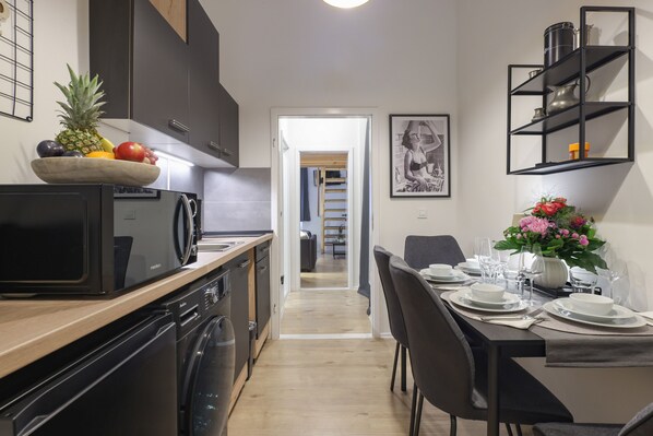 A modern kitchen with a wooden countertop, a built-in dishwasher, and a microwave. There is a dining area with a table set for two, a vase with flowers, and a black shelf unit above the table.