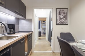 Une cuisine moderne avec des meubles de couleur sombre, une machine à café et un évier. On aperçoit, à travers une porte ouverte, un coin repas avec une table et des chaises.