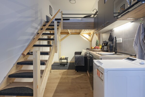 A compact kitchen with a built-in washing machine, a wooden staircase, and a small seating area.