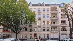 Um edifício residencial com vários andares, varandas, carros estacionados e uma árvore em primeiro plano.