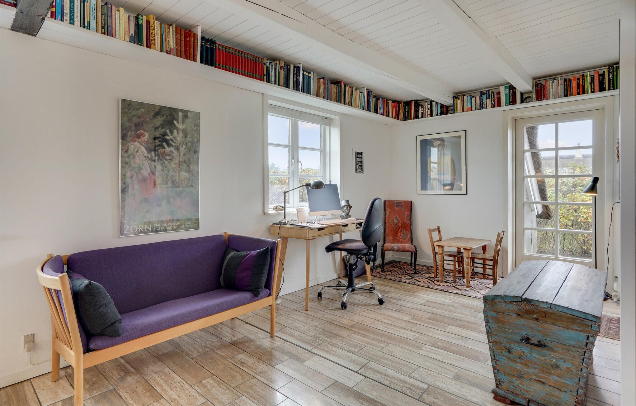 A room with a purple sofa, a wooden desk with a computer, a chair, a dining table with chairs, and a bookshelf filled with books.
