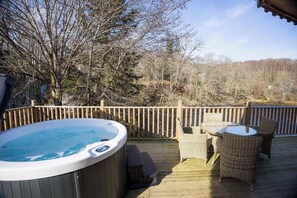 A wooden deck with a hot tub, wicker chairs, and a view of trees and hills. - Otters Holt Circular Riverside Retreat with Hot Tub (Killin)