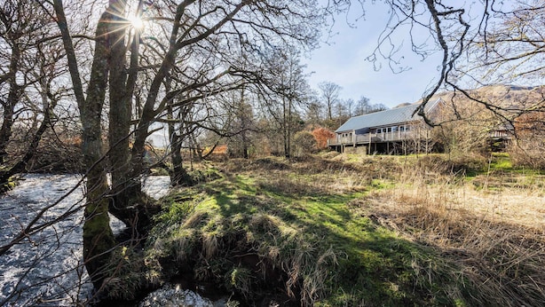 A house with a deck, surrounded by trees and a stream. - Cosy Riverside Escape with Hot Tub | Garth Cottage (Killin)