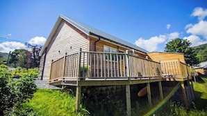 A wooden cabin with a balcony, surrounded by greenery. - Cosy Riverside Escape with Hot Tub | Garth Cottage (Killin)