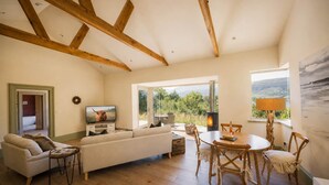 A living room with a sofa, TV, wooden beams, a fireplace, and a view of the outdoors. - The Tractor Shed- Elevated Hot Tub with Loch Views (Acharn)