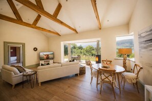 A living room with a sofa, TV, wooden beams, a fireplace, and a view of the outdoors.
