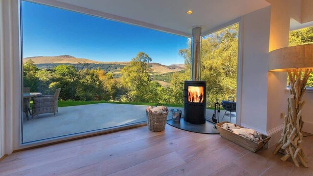 A modern interior with a large glass door leading to a deck, a wood-burning stove, wicker furniture, and a rustic decor piece. - The Tractor Shed- Elevated Hot Tub with Loch Views (Acharn)