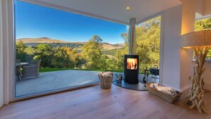 A modern interior with a large glass door leading to a deck, a wood-burning stove, wicker furniture, and a rustic decor piece. - The Tractor Shed- Elevated Hot Tub with Loch Views (Acharn)