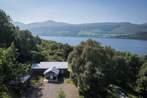 Una casa con techo metálico, rodeada de árboles frondosos, con vista a un lago y montañas a lo lejos.