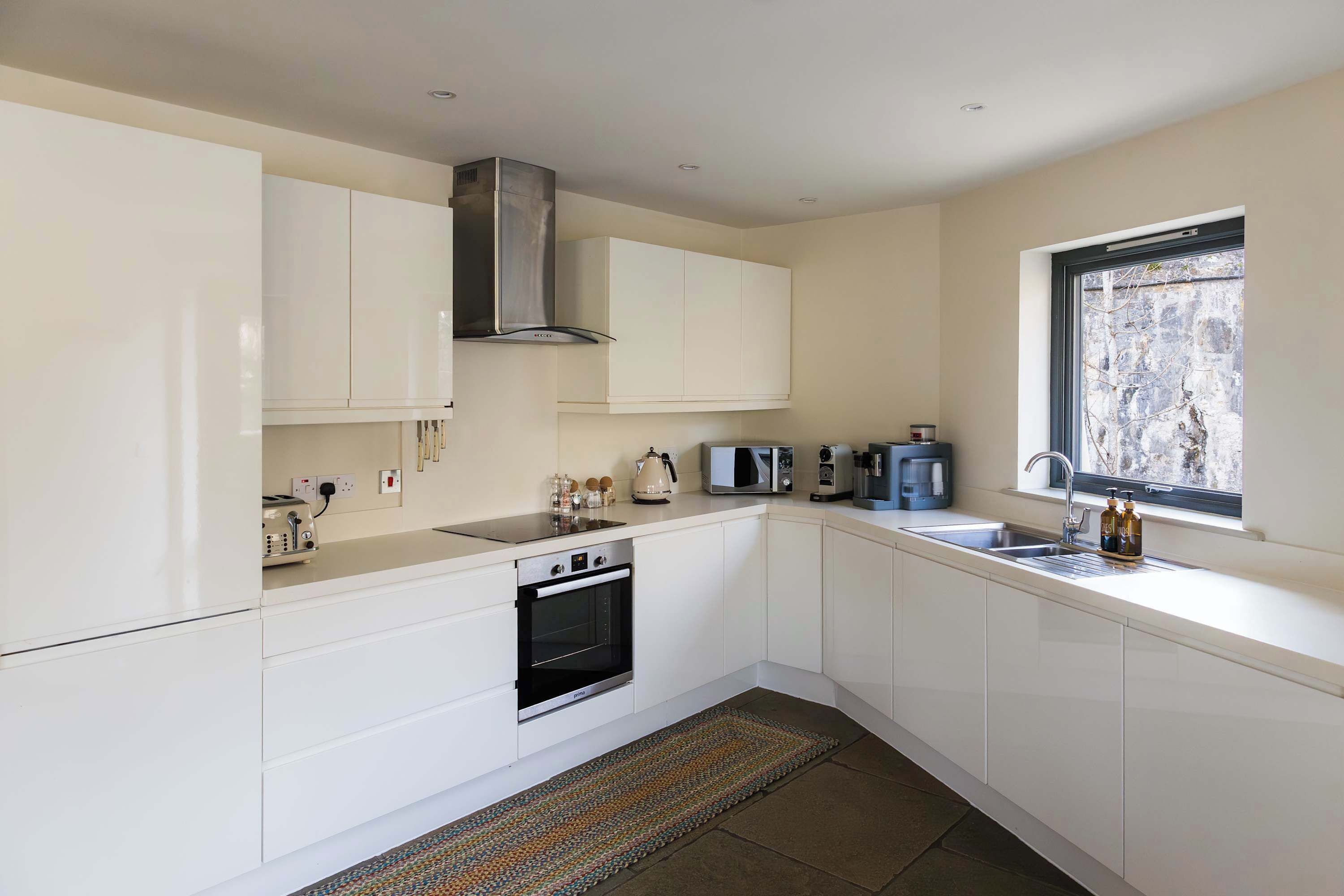 A modern kitchen with white cabinetry, a built-in oven, a microwave, a kettle, and a window with a view.