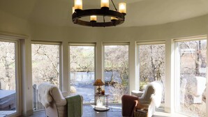 A room with a stone floor, a chandelier, two chairs with throws, a small table with a lamp, and large windows with a view of trees and a body of water. - Luxury Riverside Lodge with Hot Tub (Sleeps 6) (Killin)