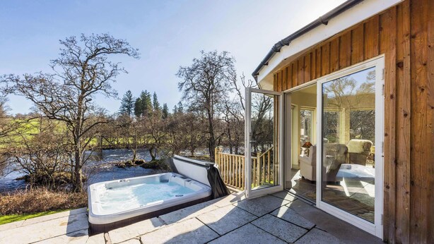 A hot tub on a patio with a wooden deck, a glass door leading to a room with a chair, and a view of a river and trees. - Luxury Riverside Lodge with Hot Tub (Sleeps 6) (Killin)