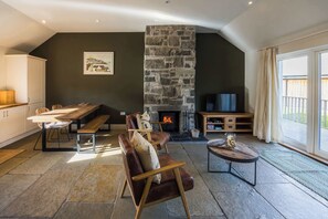 A cozy living room with a stone fireplace, a dining area with a bench, and a TV. - Ghillie's Cottage on the River Lochay (Killin)