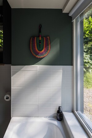 A bathroom with a white bathtub, a window with a view of greenery, and a colorful woven item on the wall. - Osprey Boathouse Over Loch Tay with Hot Tub (Acharn)