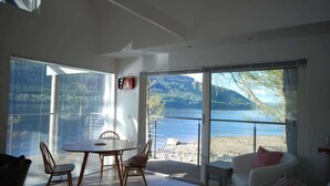A modern living room with a large sliding glass door leading to a balcony overlooking a lake and mountains. - Dipper Boathouse Secluded Over-Water Retreat (Acharn)
