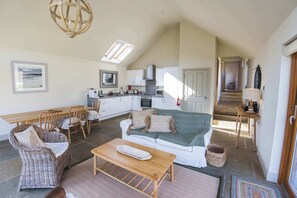 A spacious living area with a sofa, coffee table, wicker chairs, and a dining table. A kitchen with white cabinets and a staircase leading to an upper level is visible. - Beautiful river fronted house with private hot tub (Killin)