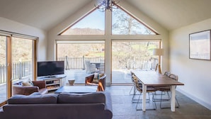A spacious living area with a large sofa, a television, and a dining table with chairs. There are large windows providing a view of the outdoors. - Riverbank- Glass-Fronted Riverside Stay -Hot Tub (Killin)