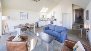 A spacious living area with a sofa, coffee table, and dining table. A kitchen with white cabinets and a staircase is visible in the background. - Riverbank- Glass-Fronted Riverside Stay -Hot Tub (Killin)
